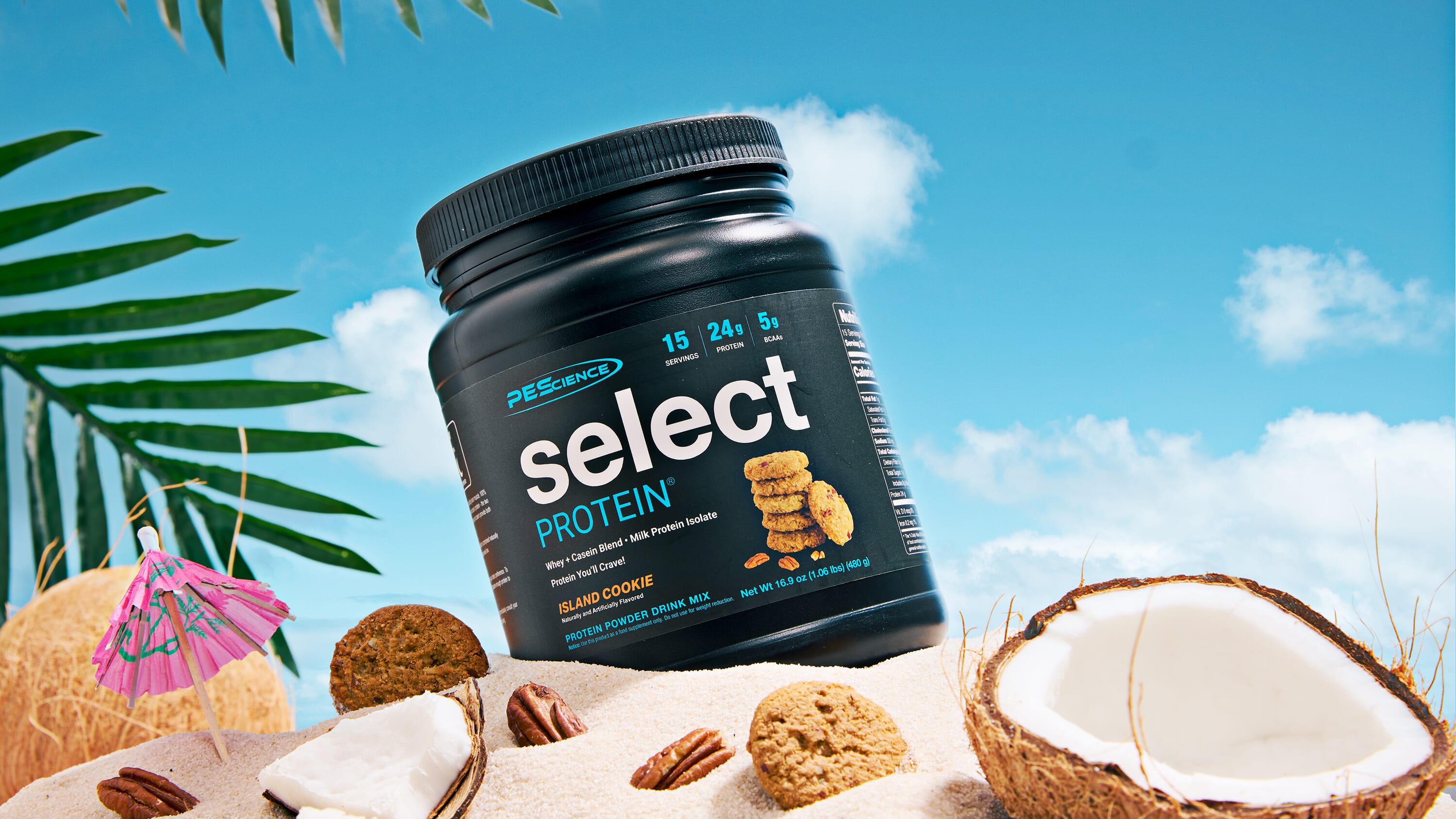 A tub of PreWorkout Select Protein in Island Chocoe flavor, styled on a sandy beach with a blue sky background, coconut halves, pecans, and tropical palm leaves.