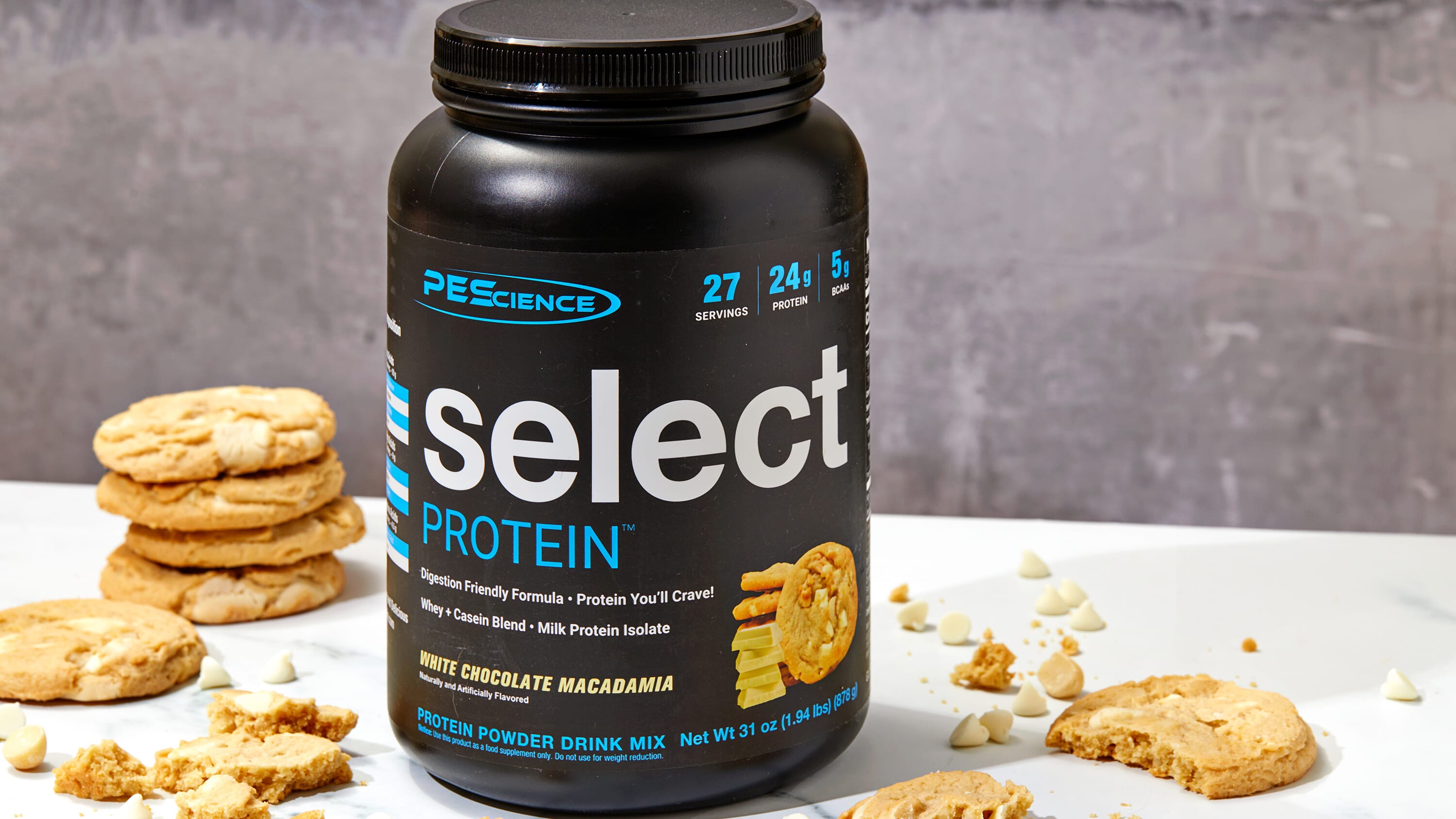 A tub of PEScience Select Protein powder in White Chocolate Macadamia flavor, styled on a white surface with cookie props against a gray textured background.